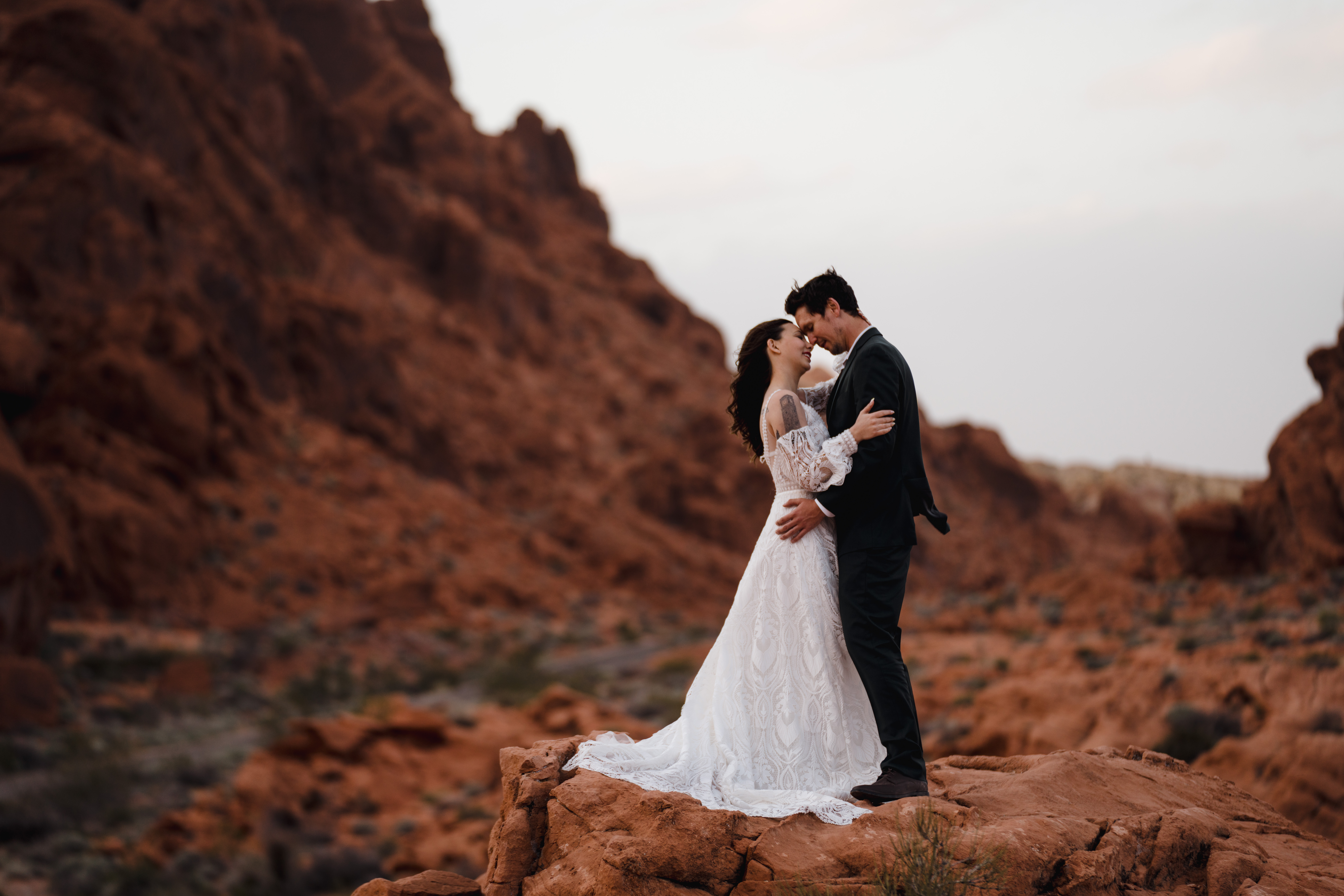 A bride and groom embrace and kiss on a rocky terrain with a backdrop of red sandstone formations, expertly captured by a wedding photographer in Las Vegas in Valley of Fire, Nevada