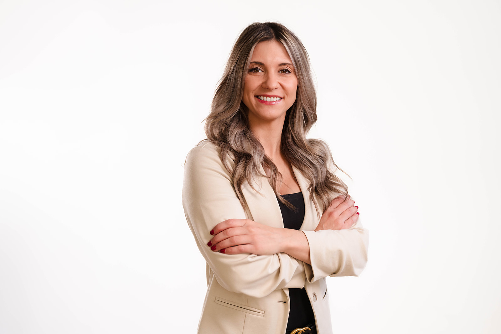 In this stunning example of studio photography, a person with long hair stands against a white background, wearing a beige blazer and black top, smiling with arms crossed. The composition captures the essence of a professional headshot photographer's work.