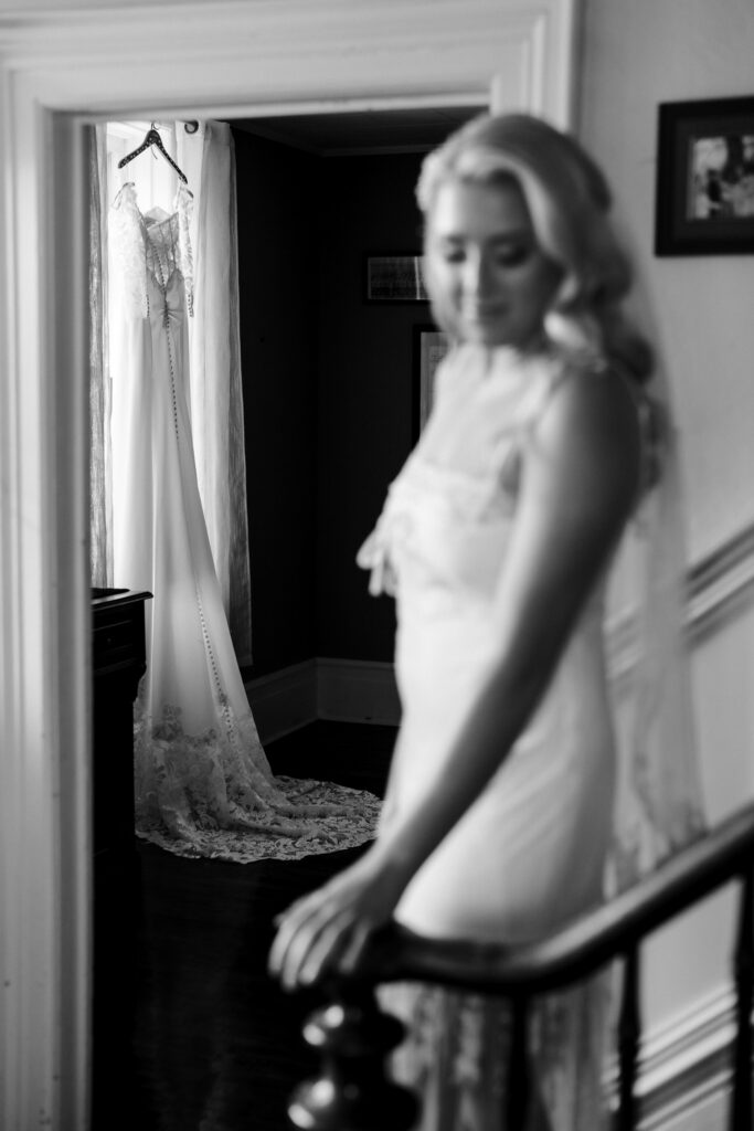Black and white portrait of Ainsley in her veil on the staircase, her wedding dress hanging in the doorway behind her
