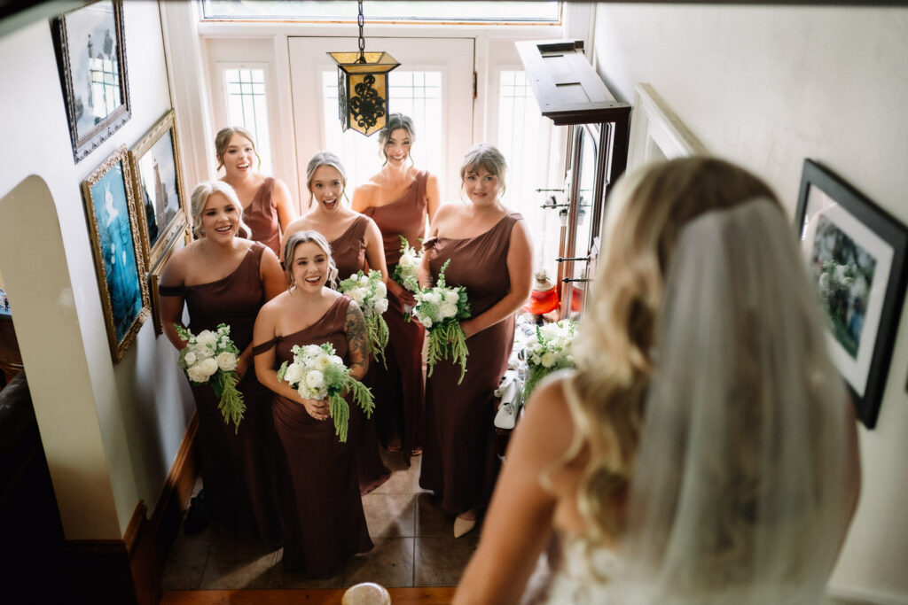 Bridesmaids in rust dresses smiling as they see Ainsley in her wedding dress and veil