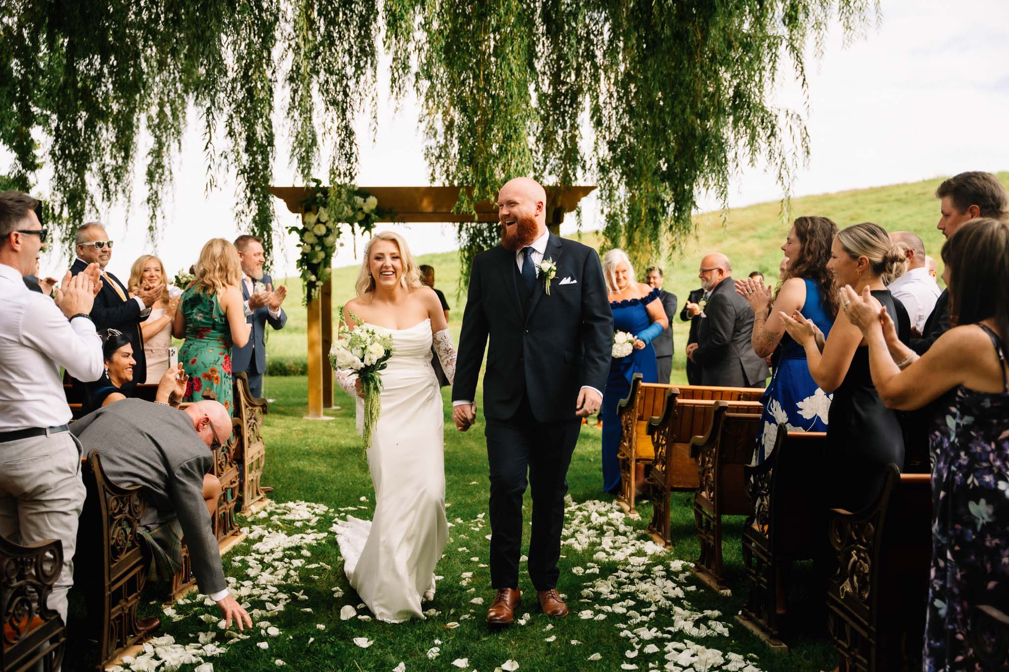 Ainsley and Rob walking back up the aisle at their Polmenna Barn wedding as guests cheer and petals cover the grass