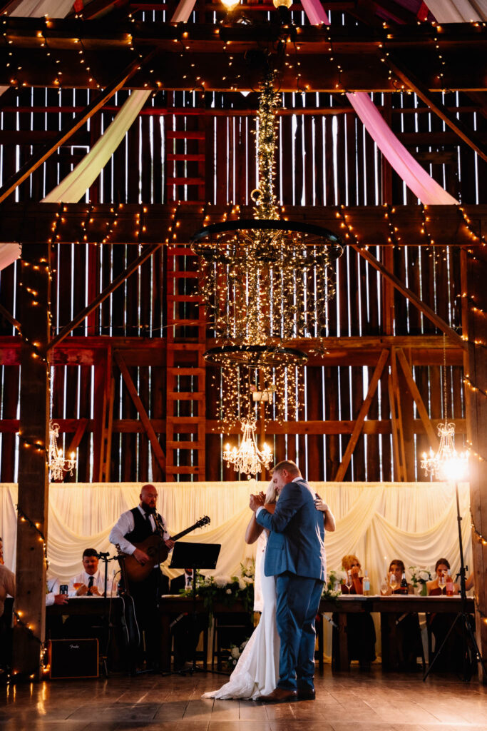 Polmenna Barn wedding reception with Ainsley slow dancing under string lights and a chandelier while a guitarist plays behind her