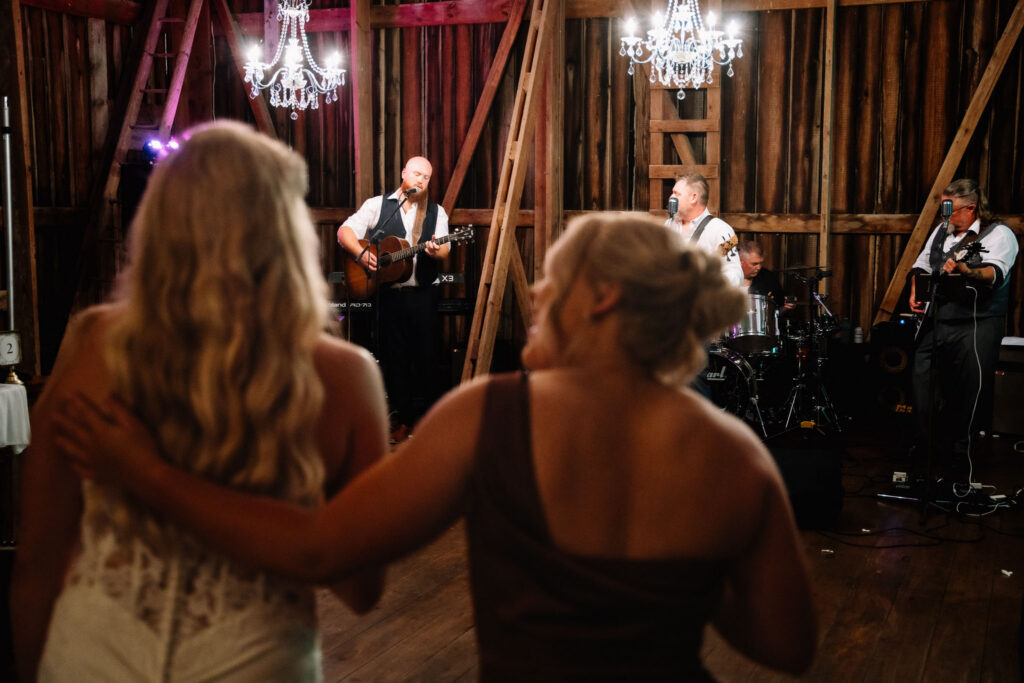 Ainsley and a bridesmaid arm in arm on the dance floor, watching the live band play in the barn