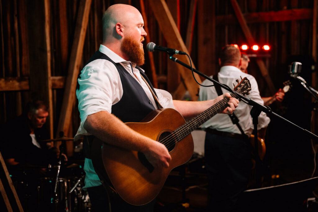 Bearded guitarist singing at the microphone on the barn stage with the band playing behind him
