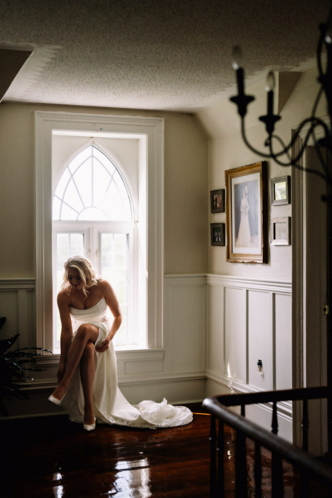 Ainsley getting ready beside a tall arched window on the morning of her Polmenna Barn wedding, her dress trailing across the hardwood floor
