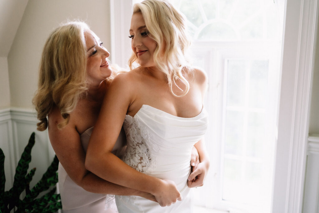 Ainsley in her wedding gown smiling as a woman hugs her from behind in front of a bright window