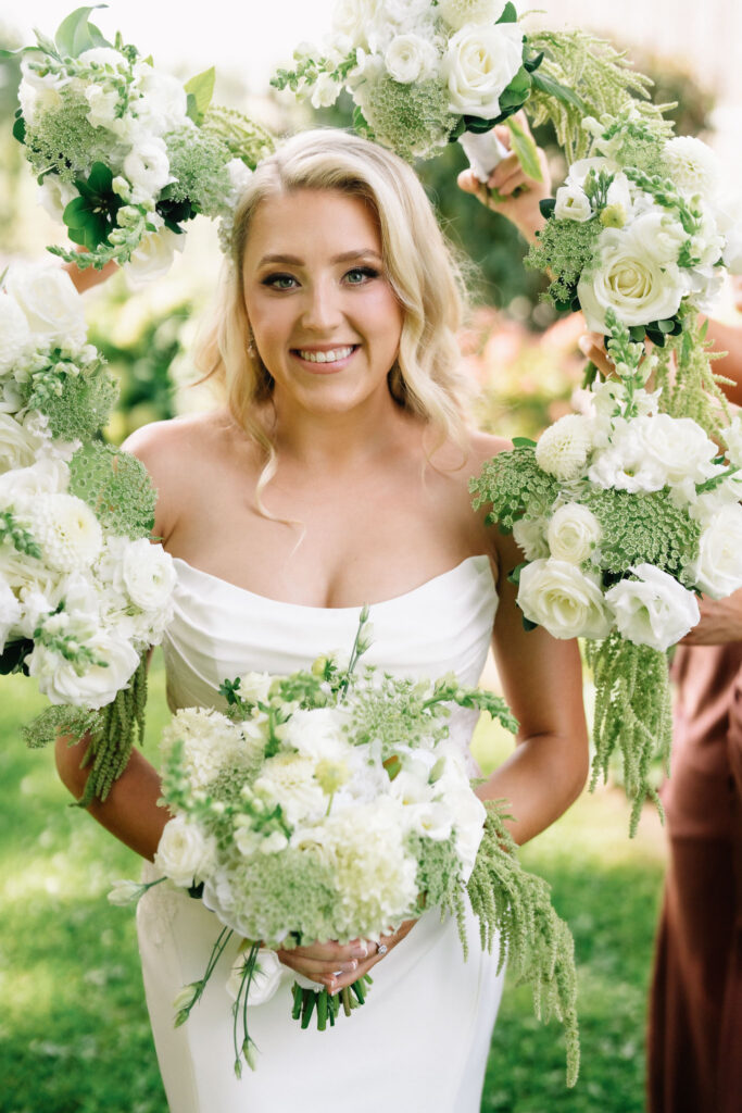 Ainsley smiling at the camera with her white and green bouquet on the day of her Polmenna Barn wedding
