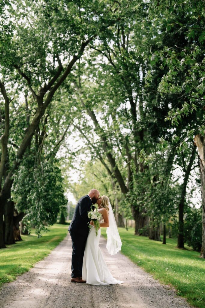 Ainsley and Rob embracing on a gravel laneway under a canopy of trees at their Polmenna Barn wedding