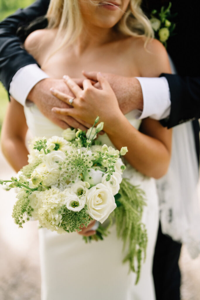 Close-up of Rob's arms around Ainsley with her wedding ring and bouquet in the frame