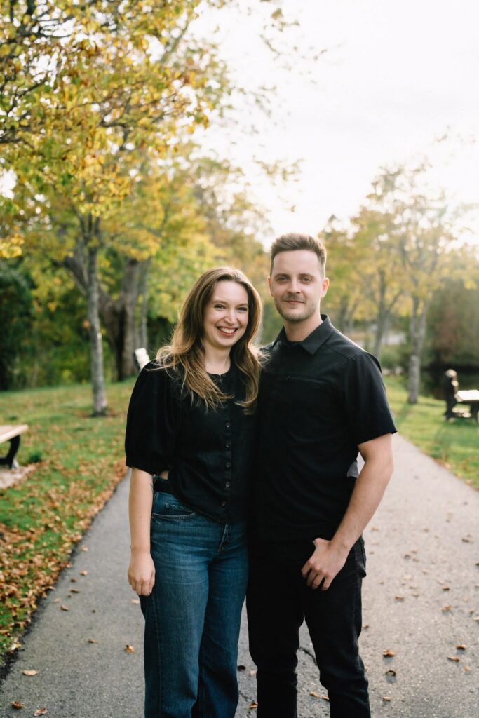 Fall engagement photo of Alexandre and Briar smiling side by side on a tree-lined path at Rockwood Park