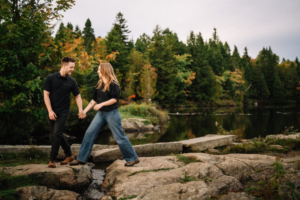 Engagement photo of Alexandre leading Briar by the hand across flat rocks at the pond, orange maples behind them