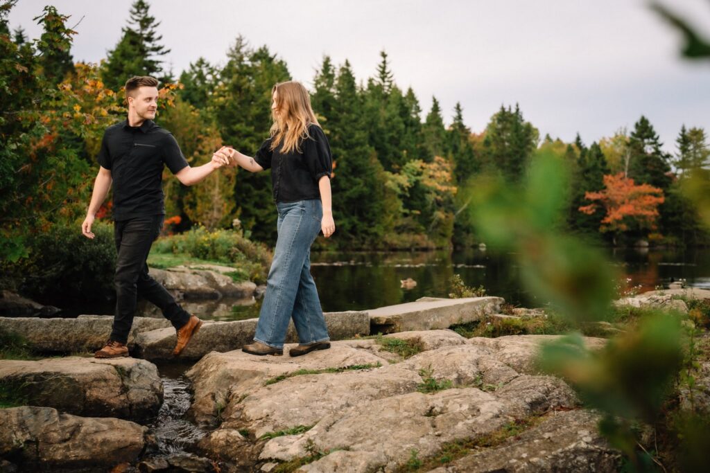 Alexandre helping Briar step across the rocks at the pond's edge with fall colour in the background
