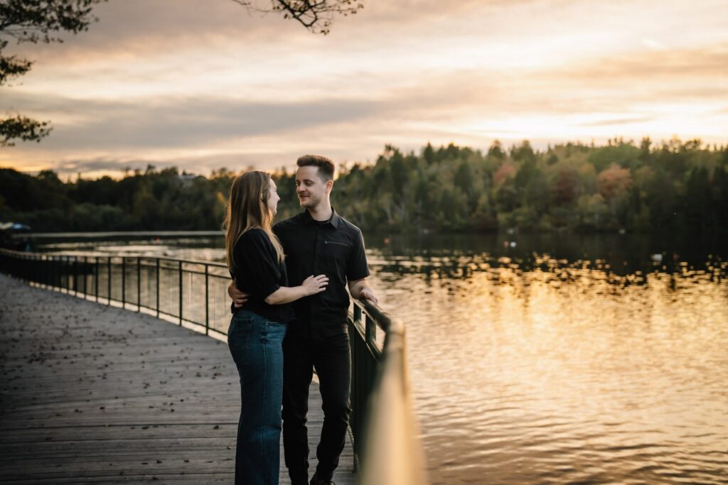 Engagement photo of Alexandre and Briar at a wooden boardwalk railing at sunset with golden light on the lake