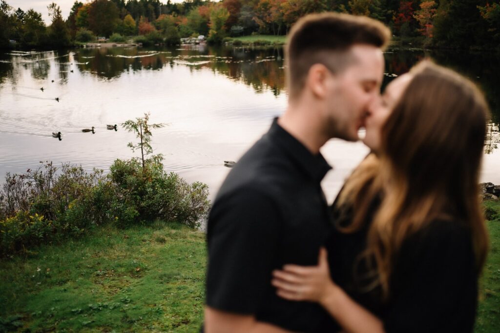 Alexandre and Briar sharing a kiss beside the pond at Rockwood Park with the water softly blurred behind them