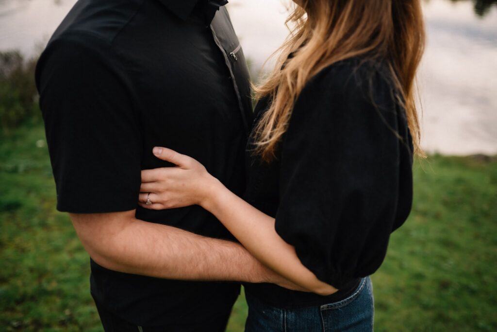 Close-up of Alexandre and Briar embracing on the grass beside the pond, Briar's arm around him
