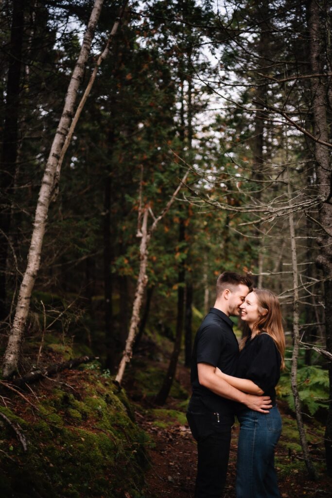 Portrait photo of Alexandre and Briar embracing at the base of tall trees on a forest trail