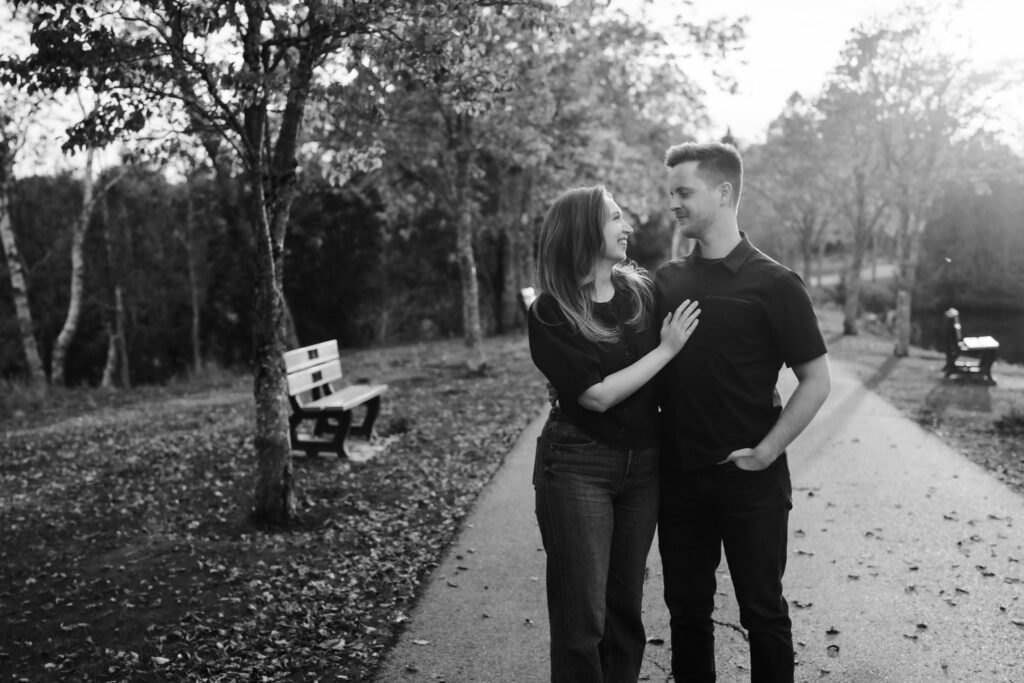 Black and white engagement photo of Briar and Alexandre embracing on a leaf-covered path beside a park bench