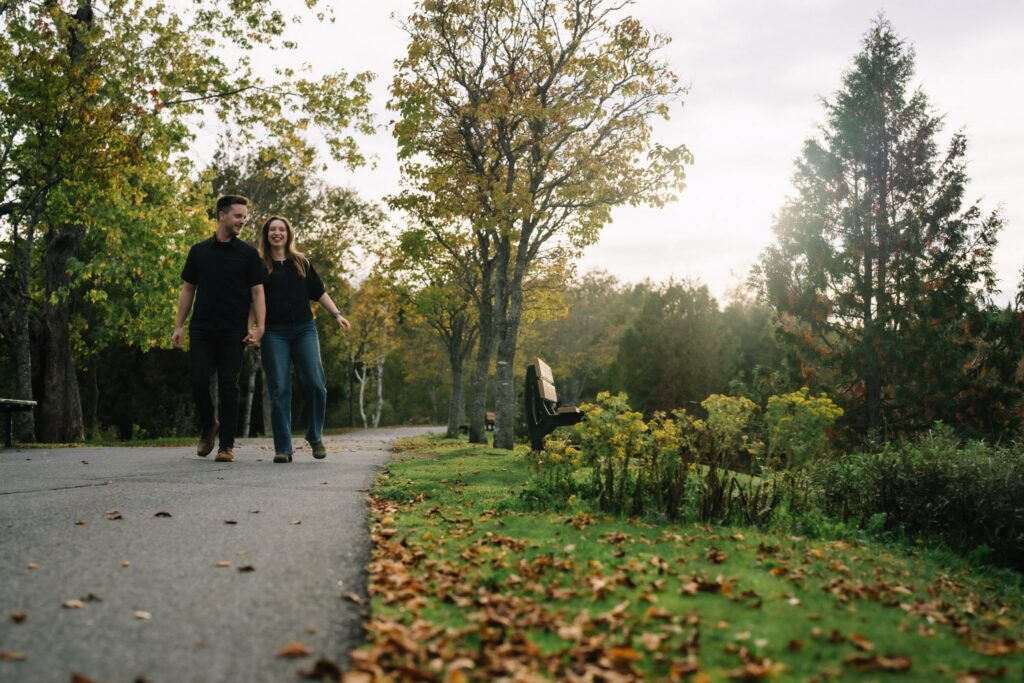 Alexandre and Briar walking away down a paved path past fall leaves and evergreens in Rockwood Park