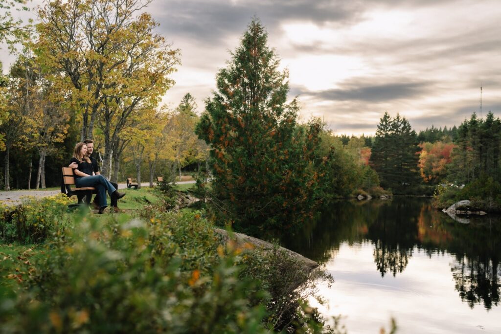 Alexandre and Briar sitting on a bench in the distance beside a calm pond reflecting evergreens and fall colour