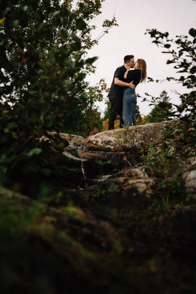 Alexandre and Briar embracing on a rocky ledge in the forest at Rockwood Park, framed by dark foliage